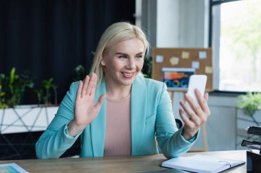 Smiling psychologist having video call on smartphone near notebook in consultation room 