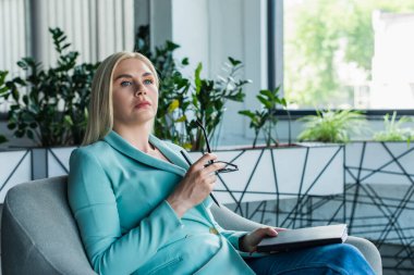 Psychologist holding notebook and eyeglasses while sitting on armchair in consultation room 
