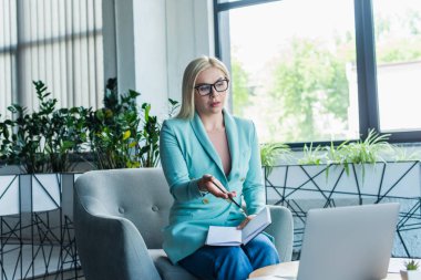 Psychologist holding notebook while having video call on laptop in consultation room 