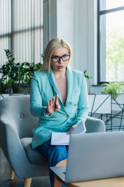 Psychologist in eyeglasses holding notebook while having video call on laptop in consulting room 