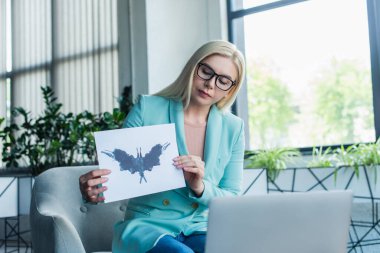 Psychologist holding Rorschach test near laptop during video call in consultation room 