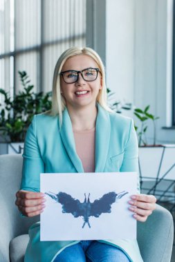 Smiling psychologist in eyeglasses holding Rorschach test in consulting room 