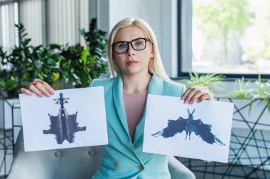 Psychologist in eyeglasses holding Rorschach test and looking at camera in consultation room 