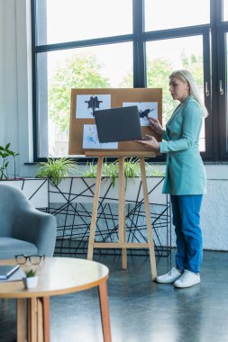 Side view of psychologist having video call on laptop near Rorschach test on board in consultation room 