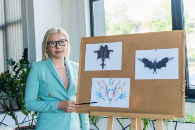Psychologist in eyeglasses pointing at Rorschach test on board in consultation room 