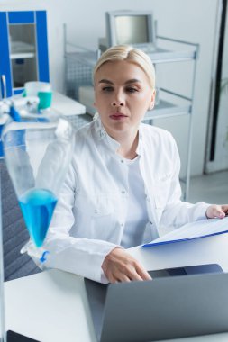 Laboratuvarda elinde dosya olan bir bilim adamı dizüstü bilgisayarın yanındaki şişeye bakıyor. 