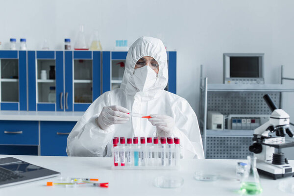 Scientist in protective goggles and suit holding test tube near laptop and microscope in lab 