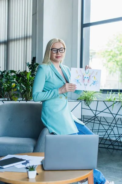 Blonde psychotherapist holding Rorschach test during video call on laptop in consultation room 
