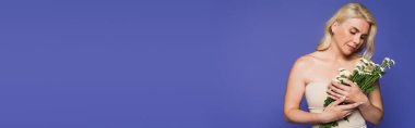 sensual woman with bare shoulders looking at blooming flowers isolated on purple, banner