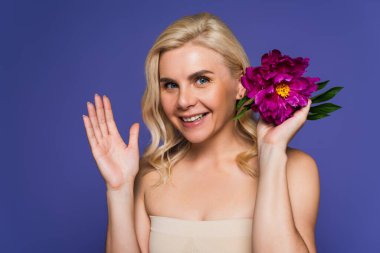 happy blonde woman with bare shoulders holding blooming flower and waving hand isolated on purple