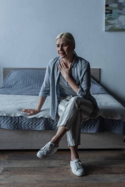 full length of upset blonde woman with menopause sitting on bed and touching chest