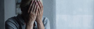 depressed blonde woman covering face behind wet window with rain drops, banner