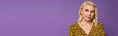 happy and blonde woman in blouse looking at camera isolated on purple, banner