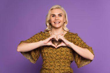 happy blonde woman showing heart sign with hands isolated on purple