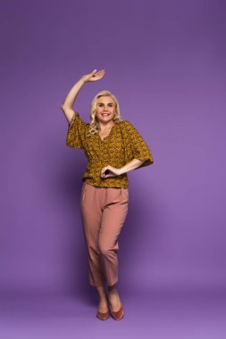 full length of joyful blonde woman in blouse having menopause and dancing on purple