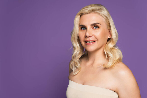 happy blonde woman with bare shoulders looking at camera isolated on purple