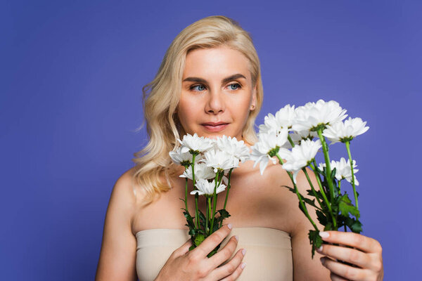 blonde woman with bare shoulders looking at blooming flowers isolated on violet