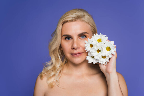 blonde woman with bare shoulders holding flowers and looking at camera isolated on violet