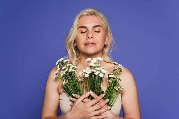 blonde woman with closed eyes smelling flowers isolated on purple