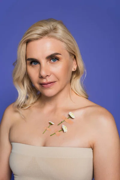 blonde woman with plasters and tiny flowers on body looking at camera isolated on purple