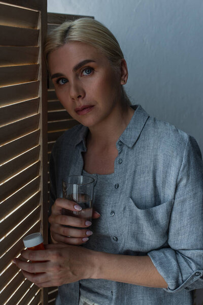 blonde woman with menopause holding glass of water near folding screen 