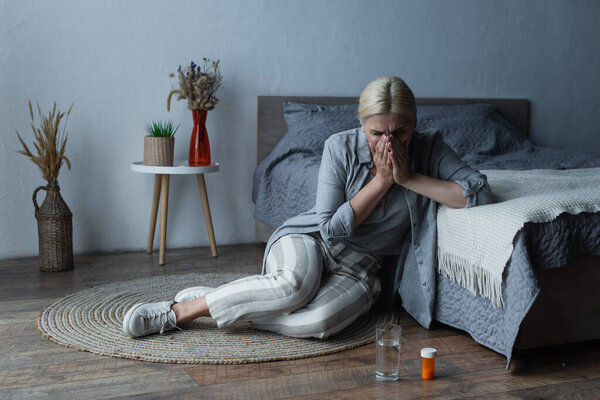 woman with menopause sitting near bed while covering face and suffering from abdominal pain 