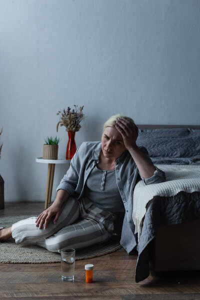 exhausted woman sitting near bed and looking at glass of water and painkillers 