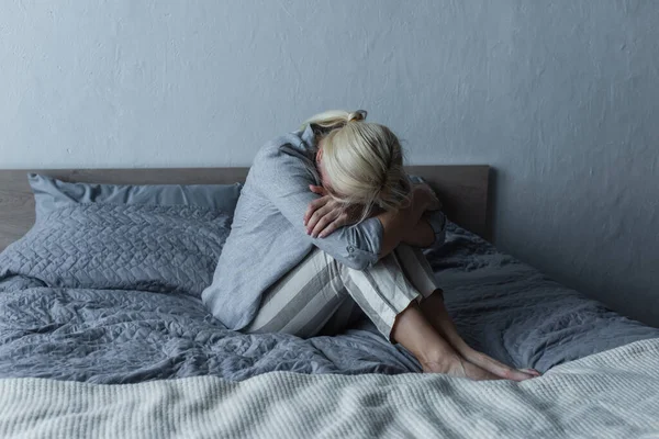 Blonde Woman Covering Face Hugging Knees Bed While Feeling Pain — 스톡 사진