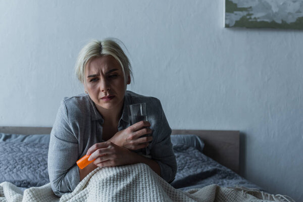 exhausted woman with climax sitting in bed while holding glass of water and painkillers 
