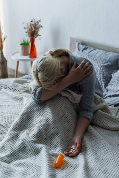blonde woman with climax covering face while sitting in bed with painkillers in hand
