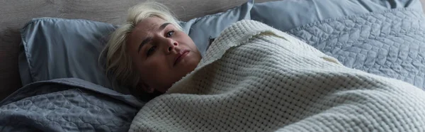 depressed blonde woman with menopause lying under blanket in bed, banner