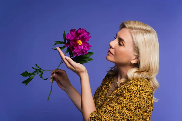 side view of blonde woman in blouse looking at purple flower with green leaves isolated on violet