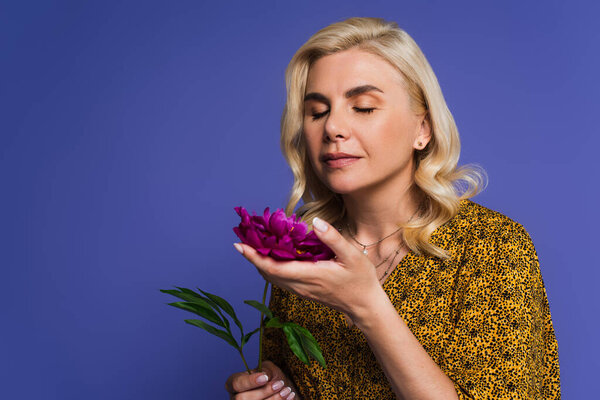 blonde woman in blouse smelling purple flower with green leaves isolated on violet