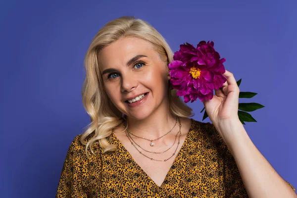 joyful woman in blouse holding purple flower with green leaves isolated on violet