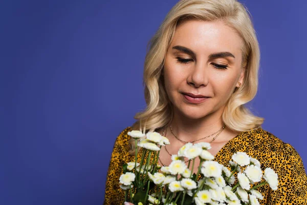 blonde woman in blouse looking at bouquet of white flowers isolated on violet