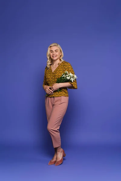 full length of happy woman in blouse holding bouquet of white flowers with green leaves on violet