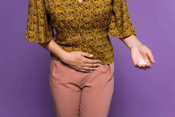 cropped view of woman with stomach ache holding pills isolated on purple