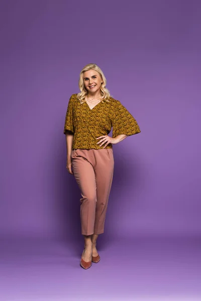 full length of blonde woman with menopause posing with hand on hip and smiling on purple