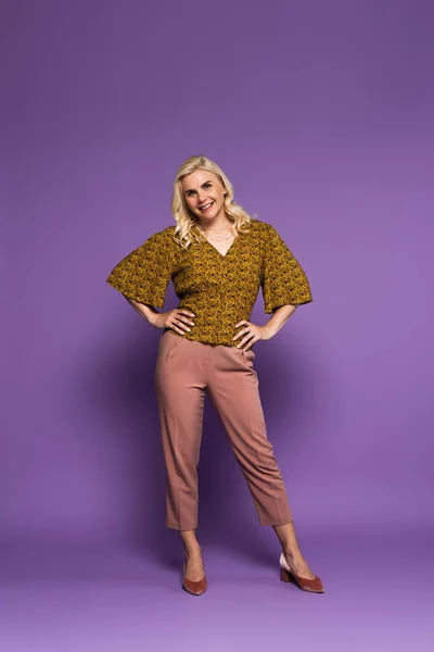 full length of blonde woman with menopause posing with hands on hips and smiling on purple