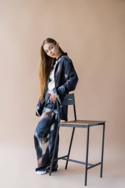 Bej arka planda sandalyenin yanında poz veren uzun saçlı bir kadın.