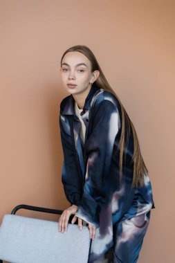Moda kıyafetleri içinde güzel bir kadın bej koltuğun yanındaki kameraya bakıyor.