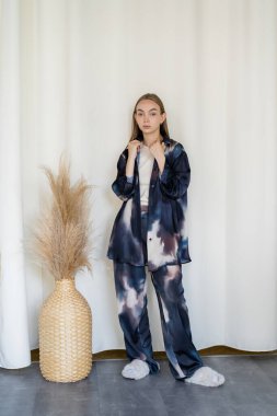 full length of woman in trendy oversize clothes and fluffy slippers near spikelets in wicker vase on white background