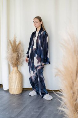 full length of woman in oversize suit and fluffy slippers near wicker vase with spikelets and white drapery