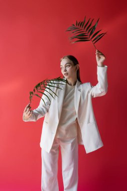 young woman in white clothes posing with green tropical leaves on crimson