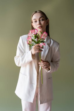 Yeşil üzerinde pembe alstromerya çiçekleri tutan beyaz ceketli genç bir kadın.