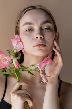 Alstromeria dalına sahip şehvetli bir kadın bej rengi tenha bir yanağa dokunuyor.