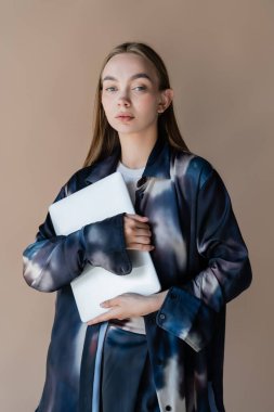 Kravatlı, renkli tişörtlü çekici bir kadın kameraya bakıyor ve elinde bej bir dizüstü bilgisayar tutuyor.