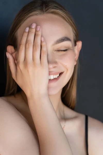 portrait of smiling woman with natural makeup obscuring face with hand isolated on black