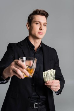 confident man holding dollar banknotes and toasting with blurred glass of whiskey isolated on grey clipart