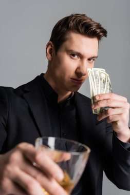 confident man showing dollar banknotes and toasting with blurred glass of whiskey isolated on grey
